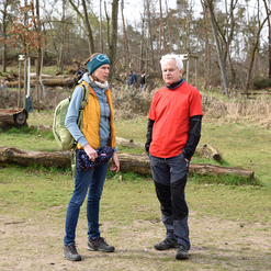 Zwei Wanderer stehen im Gespräch auf einem grünen Waldweg, umgeben von Bäumen und umgestürzten Stämmen.