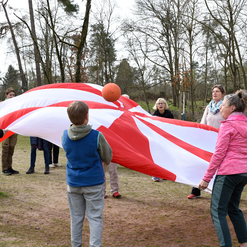 Gruppe von Menschen spielt mit einem großen, rot-weißen Fallschirm und einem Ball auf einer Wiese im Freien.
