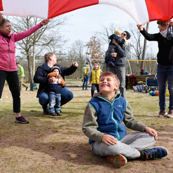 Kinder und Erwachsene spielen mit einem bunten Fallschirm im Freien, während ein Junge lächelnd sitzt.