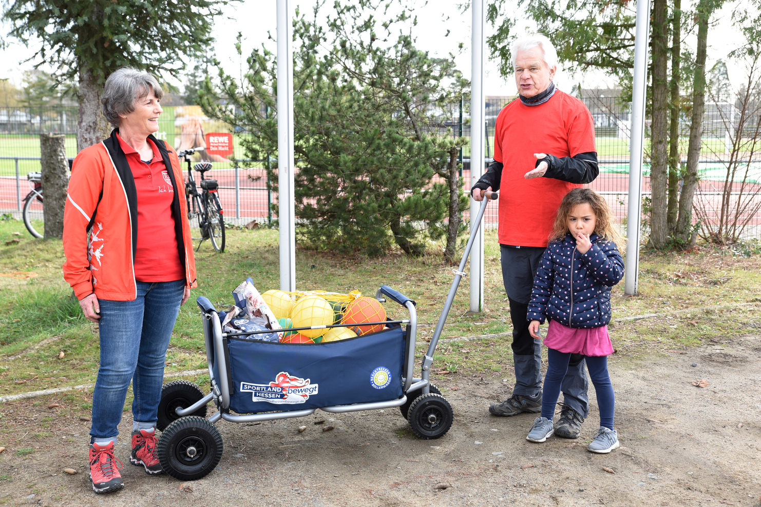 Zwei Erwachsene und ein Kind stehen an einem Sportplatz neben einem Wagen mit verschiedenen Sportbällen.