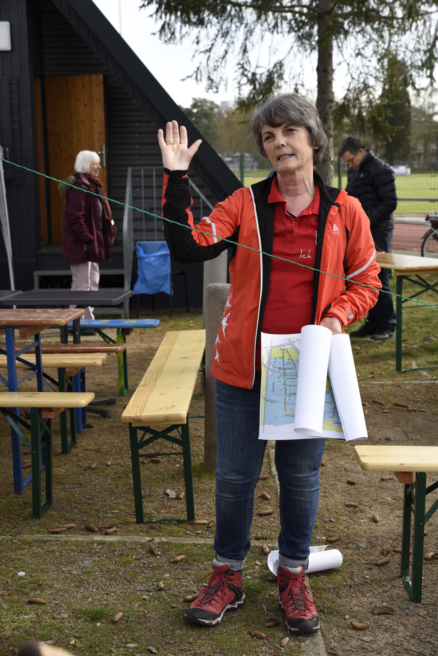 Frau mit Karten in der Hand, die im Freien spricht, während im Hintergrund Tische und andere Personen zu sehen sind.