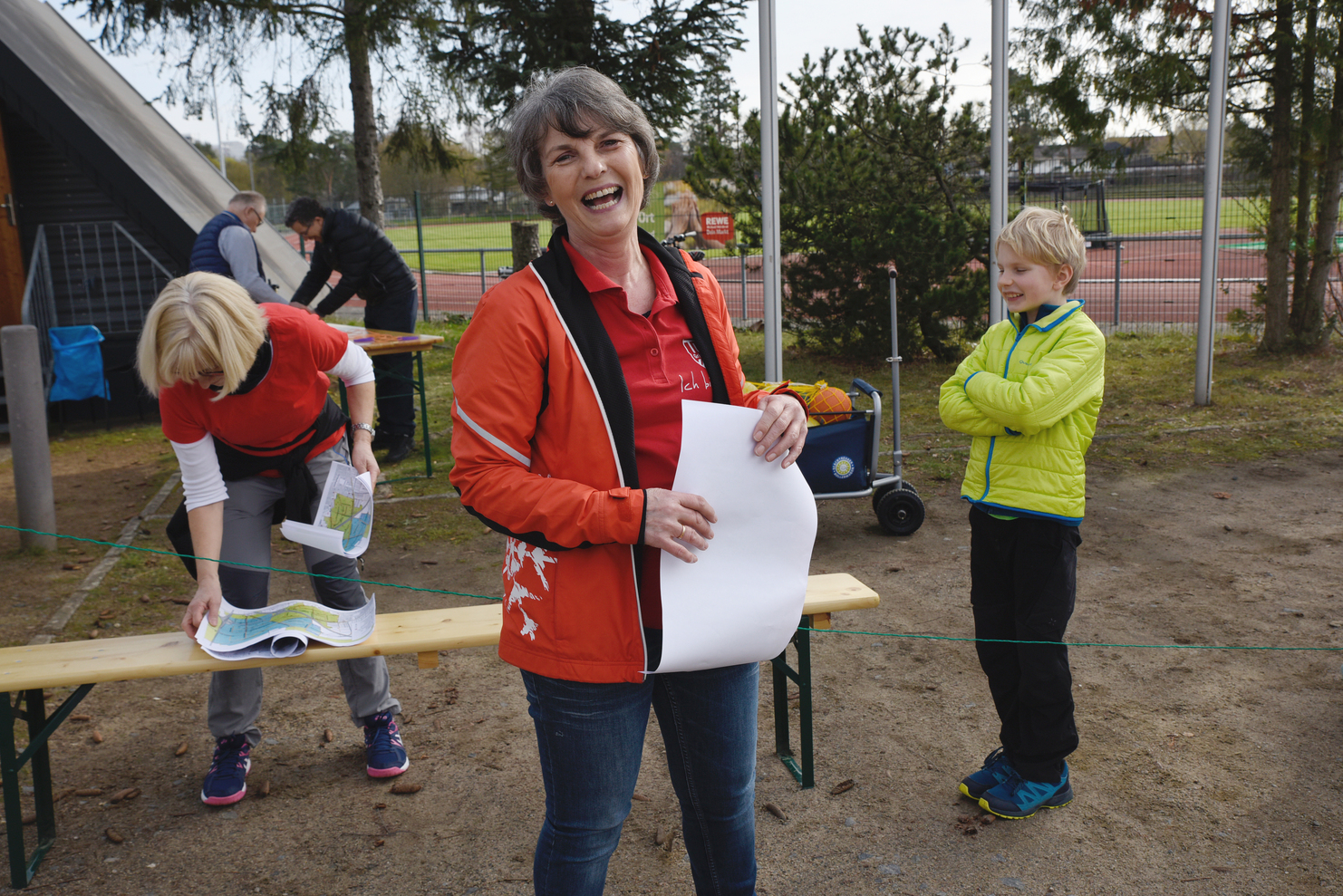 Lachende Frau in roter Jacke hält ein Blatt Papier, während ein Kind in grüner Jacke daneben steht. Andere Personen im Hintergrund.