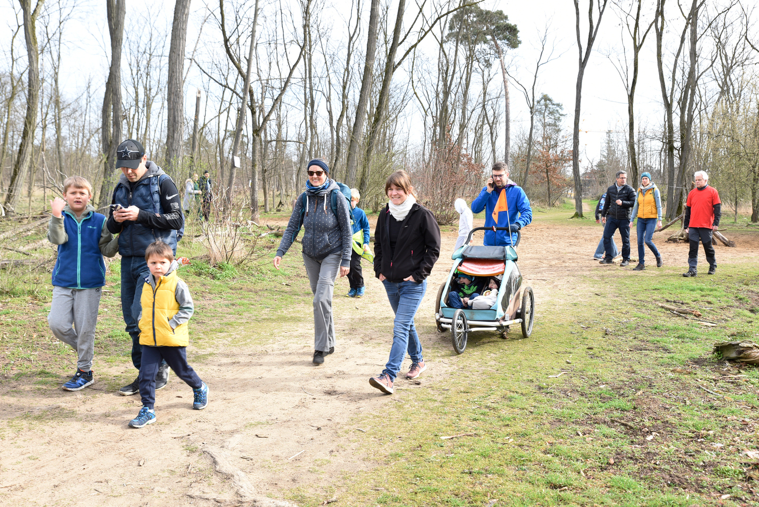 Gruppe von Erwachsenen und Kindern spaziert auf einem schmalen Weg durch einen bewaldeten Park im Frühling.