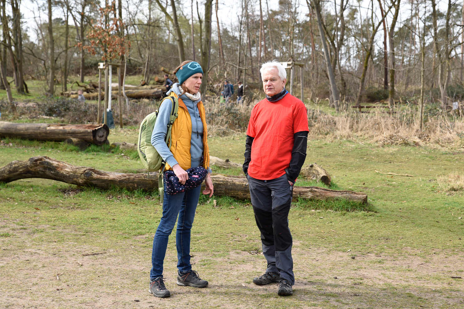 Zwei Wanderer stehen im Gespräch auf einem grünen Waldweg, umgeben von Bäumen und umgestürzten Stämmen.