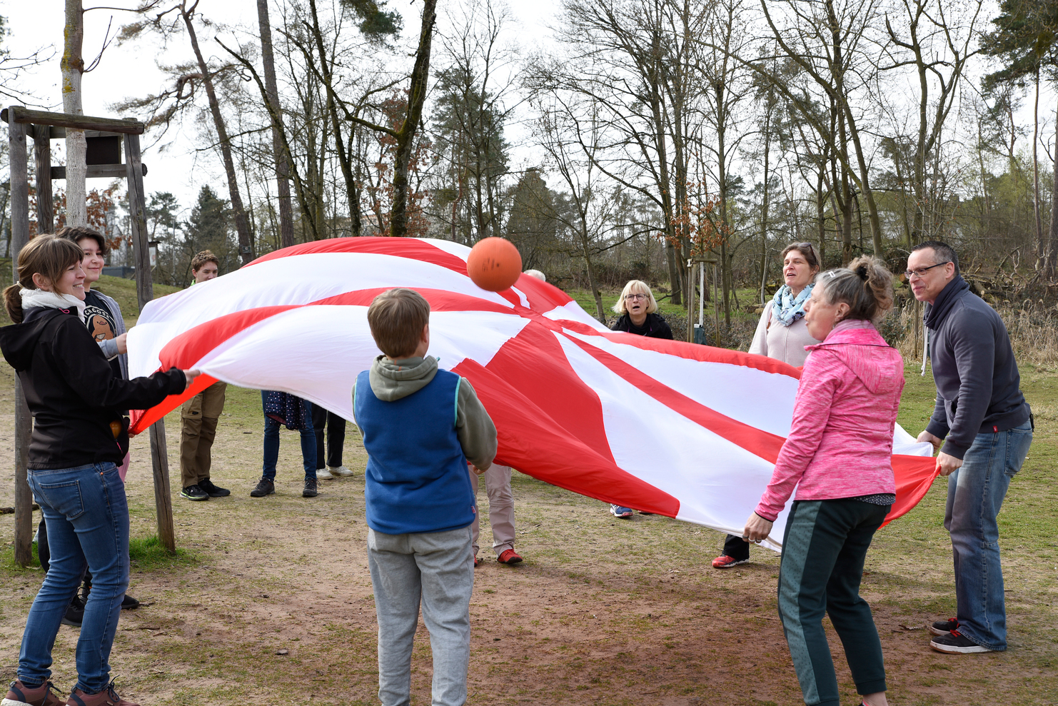 Gruppe von Menschen spielt mit einem großen, rot-weißen Fallschirm und einem Ball auf einer Wiese im Freien.