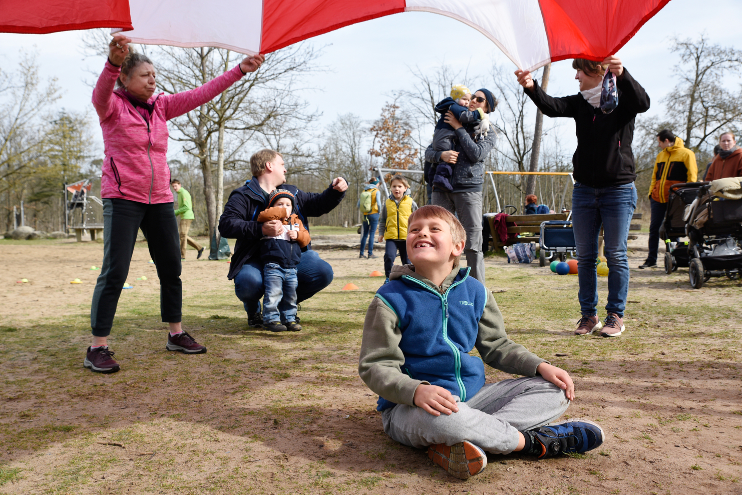 Kinder und Erwachsene spielen mit einem bunten Fallschirm im Freien, während ein Junge lächelnd sitzt.
