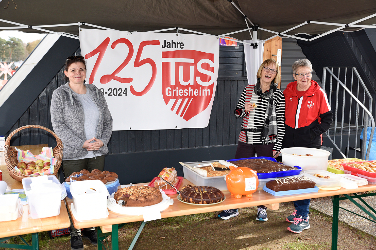Drei Frauen stehen an einem Tisch mit Kuchen und Snacks, im Hintergrund ein Banner zum 125-jährigen Jubiläum des TuS Griesheim.