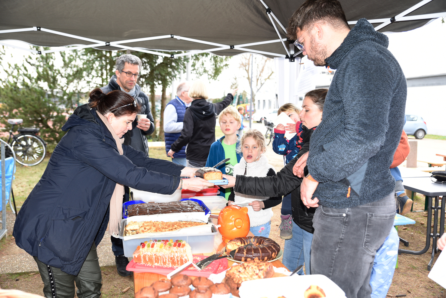 Lebhafte Szene an einem Stand mit verschiedenen Kuchen und Gebäck, während Erwachsene und Kinder miteinander interagieren.