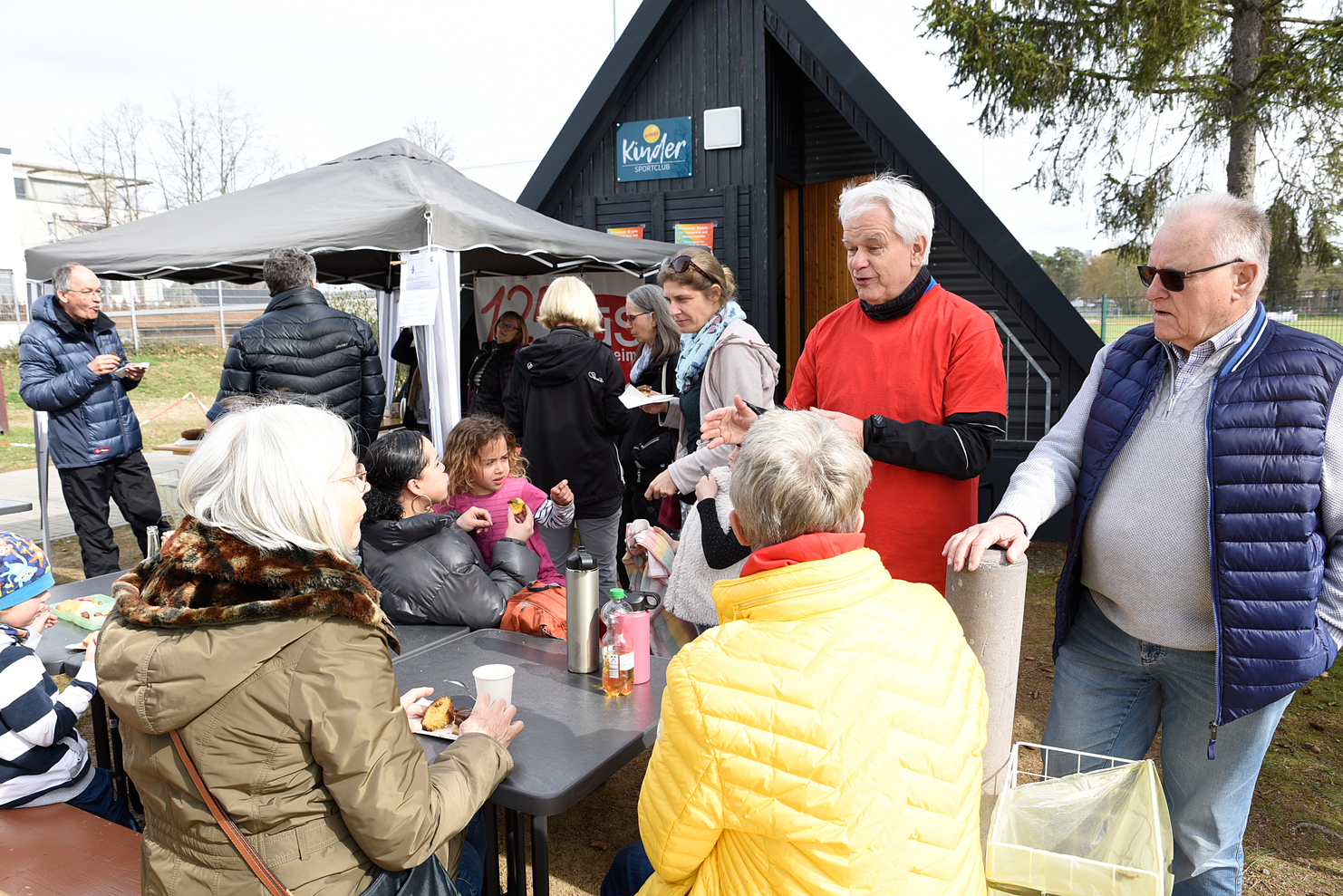 Gruppierung von Menschen am Tisch, die snacks genießen und sich unterhalten, vor einem Sportclubgebäude im Freien.