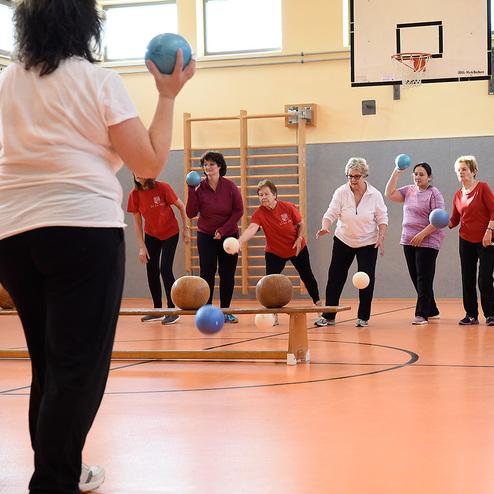 Gruppe von Frauen, die in einer Sporthalle mit bunten Bällen und Holzkugeln Übungen durchführen.
