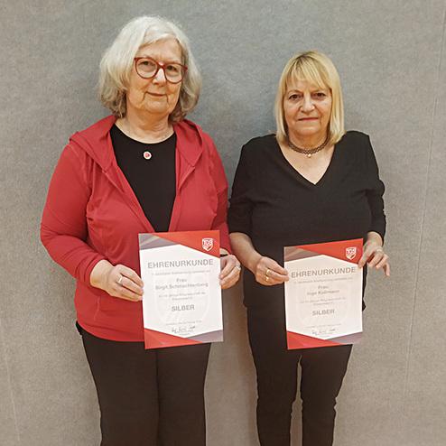 Zwei Frauen halten Urkunden f&uuml;r eine silberne Auszeichnung in einem sportlichen Rahmen, vor einer grauen Wand.