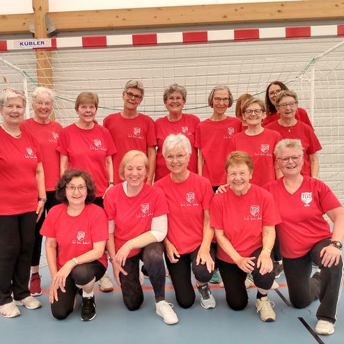 Gruppe von zwölf Frauen in roten T-Shirts vor einem Handballtor in einer Sporthalle.