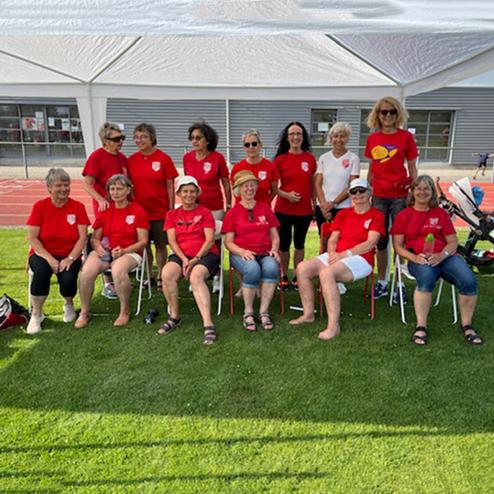 Gruppenaufnahme von 14 Frauen in roten T-Shirts, sitzend und stehend auf einer Rasenfläche vor sportlicher Kulisse.