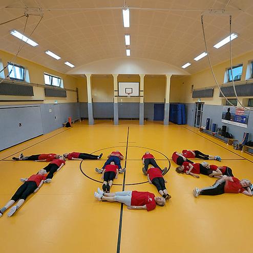 Gruppe von Sportlern in roten T-Shirts liegt auf einer orangefarbenen Sporthalle und bildet eine Zahl auf dem Boden.