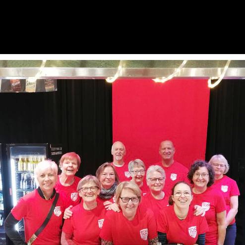 Gruppe von 14 Frauen in roten T-Shirts, lächelnd vor rotem Hintergrund in einem Veranstaltungsort.