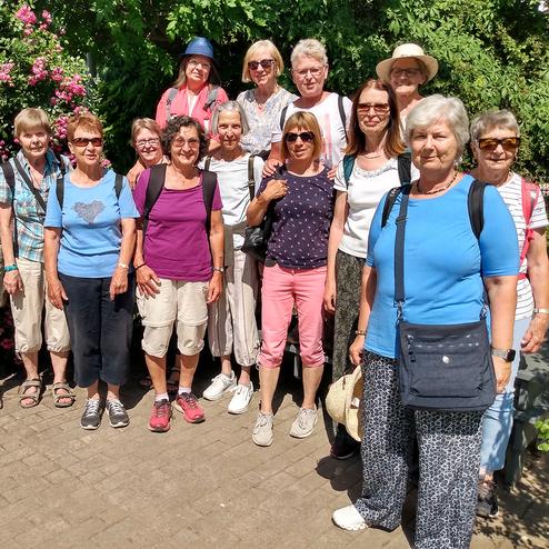 Gruppe von 15 Frauen posiert vor pink blühenden Pflanzen in einem sonnigen Garten. Alle lächeln zur Kamera.