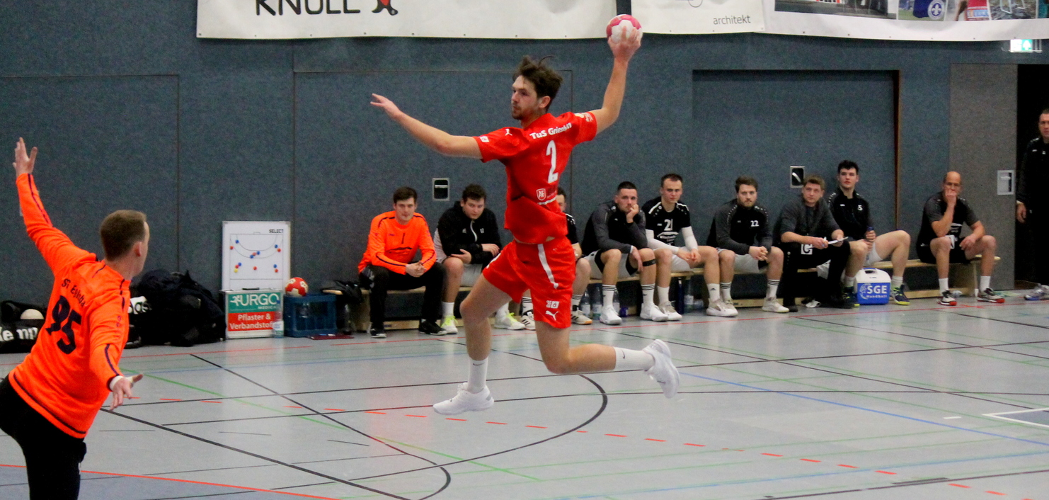 Spielszene im Hallenhandball: Ein Spieler springt und wirft den Ball in der Luft, während Teamkollegen auf der Bank zuschauen.