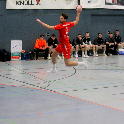 Spielszene im Hallenhandball: Ein Spieler springt und wirft den Ball in der Luft, während Teamkollegen auf der Bank zuschauen.