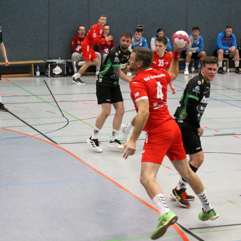 Spielszene im Handball mit Spielern in roten und schwarzen Trikots, während ein Spieler den Ball wirft. Zuschauer im Hintergrund.