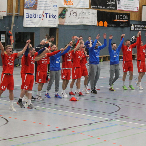 Feiernde Handballmannschaft in roten und blauen Trikots hebt die Arme nach einem Sieg auf dem Spielfeld.
