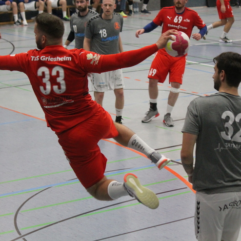 Spieler in rotem Trikot springt nach vorne, um einen Handball zu werfen, während weitere Spieler auf dem Feld stehen.