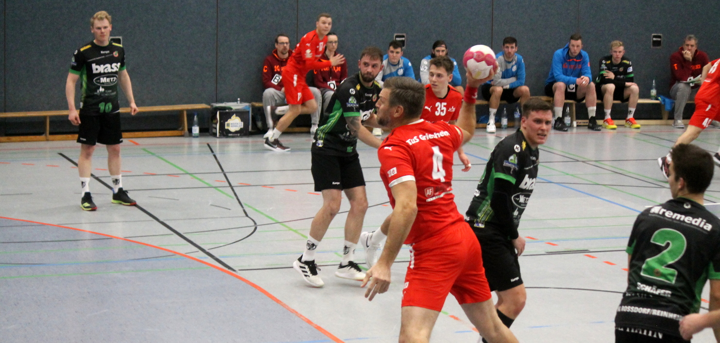 Spielszene im Handball mit Spielern in roten und schwarzen Trikots, während ein Spieler den Ball wirft. Zuschauer im Hintergrund.