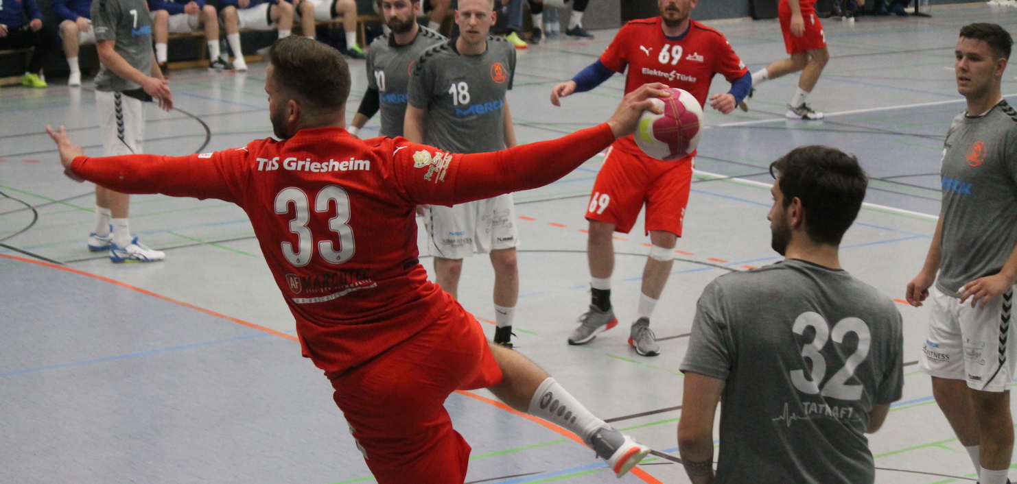 Spieler in rotem Trikot springt nach vorne, um einen Handball zu werfen, während weitere Spieler auf dem Feld stehen.