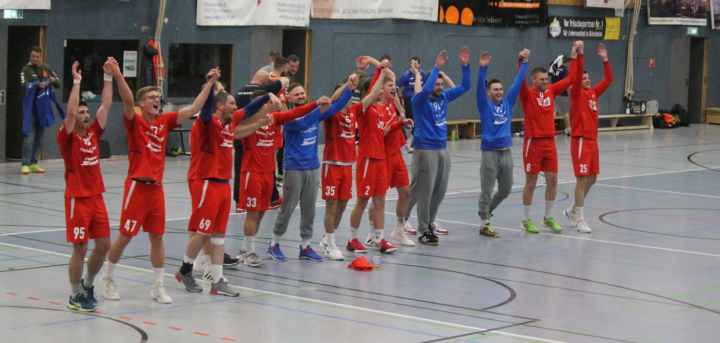 Feiernde Handballmannschaft in roten und blauen Trikots hebt die Arme nach einem Sieg auf dem Spielfeld.