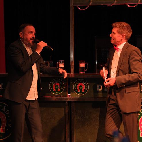 Zwei Männer in formeller Kleidung stehen an einer Bar, einer spricht ins Mikrofon. Gläser mit Bier sind sichtbar.