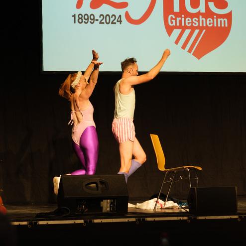 Zwei humorvoll gekleidete Personen mit Accessoires führen einen Tanz auf einer Bühne vor einem Publikum auf.