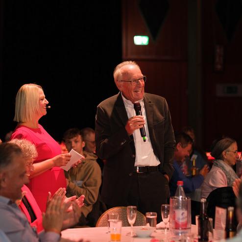 Zwei Personen sprechen auf einer Veranstaltung: Eine Frau in einem pinken Kleid und ein Mann am Mikrofon, Publikum im Hintergrund.