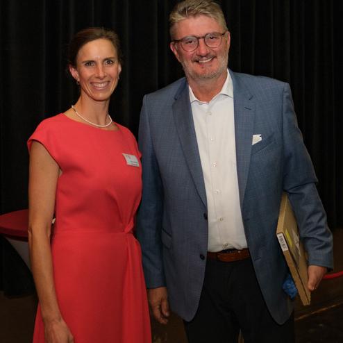 Zwei Personen lächeln in formeller Kleidung vor einem dunklen Vorhang. Die Frau trägt ein rotes Kleid, der Mann einen blauen Anzug.