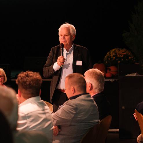 Mann mit Mikrofon spricht vor einer Gruppe von Zuhörern in einem festlichen Saal. Im Hintergrund sind Pflanzen und Dekoration sichtbar.