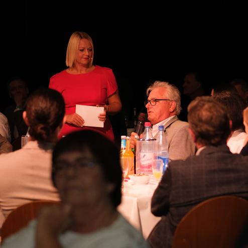 Frau in pinkem Kleid spricht vor Tischrunde, während ein Mann im grauen Anzug mit Mikrofon antwortet.