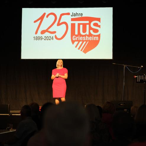 Eine Frau in pinkem Kleid steht auf der Bühne vor einem Banner zum 125-jährigen Bestehen von TuS Griesheim.