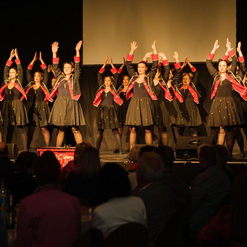 Tanzgruppe in festlicher Kleidung führt auf einer Bühne eine choreografierte Darbietung auf.