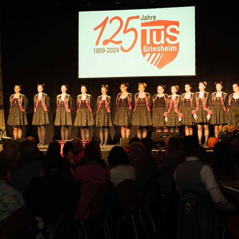 Tanzgruppe in festlicher Kleidung steht auf einer Bühne, Publikum applaudiert. Im Hintergrund das Logo „125 Jahre TuS Griesheim“.