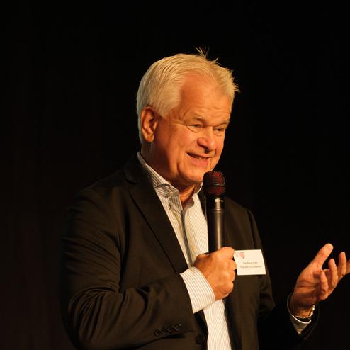 Mann mit Mikrofon hält eine Rede auf einer Bühne, trägt einen schwarzen Blazer und lächelt.