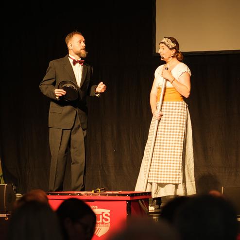 Zwei Darsteller auf einer Bühne: Mann im Anzug mit Zylinder und Frau im historischen Kleid sprechen miteinander.