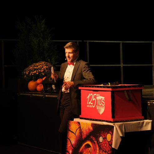 Ein Mann in einem Anzug mit Fliege spricht an einem mikrofonbestückten Tisch, umgeben von Dekorationen für eine Feier.