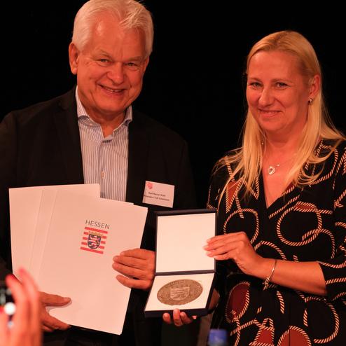 Zwei Personen stehen auf einer Bühne und halten gemeinsam eine Medaille sowie Dokumente mit dem Aufdruck „HESSEN“.
