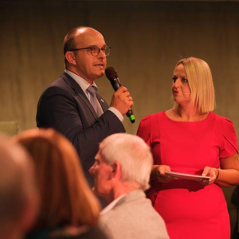 Mann mit Mikrofon spricht, während eine Frau in rotem Kleid notizen hält. Veranstaltung im Hintergrund.