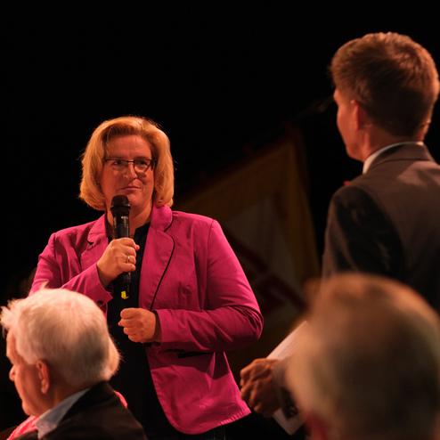 Frau in pinkem Blazer hält Mikrofon, spricht mit einem Mann auf einer Bühne; Publikum im Hintergrund.