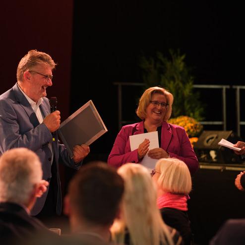 Zwei lächelnde Personen auf einer Bühne stehen zusammen, während eine der Personen ein Dokument hält.