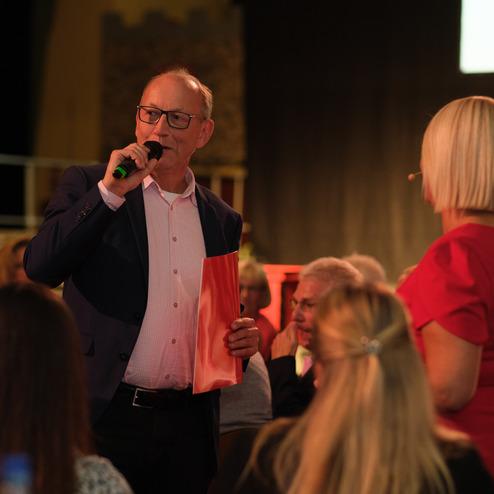 Mann mit Mikrofon spricht auf einer Veranstaltung, während eine Frau ihm aufmerksam zuhört. Publikum im Hintergrund.