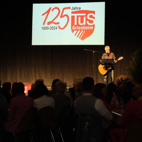 Mann mit Gitarre steht auf einer Bühne, während im Hintergrund das Logo „125 Jahre TuS Griesheim“ prangt.