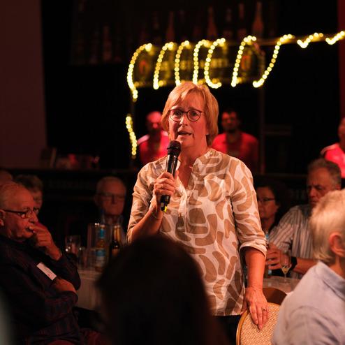 Frau mit Brille spricht mit Mikrofon in der Hand vor einem Publikum in einem festlichen Rahmen.