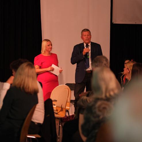 Zwei Personen sprechen auf einer Bühne, während die Zuhörer in einem festlichen Saal aufmerksam lauschen.