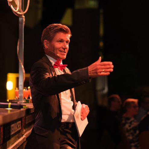 Ein Mann in Anzug mit roter Fliege spricht auf einer Veranstaltung und gestikuliert vor einem Publikum.