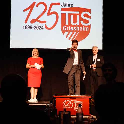 Drei Personen auf einer Bühne bei einer Veranstaltung zum 125-jährigen Jubiläum von TuS Griesheim, mit einem Logo im Hintergrund.
